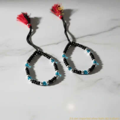 Product Description Vibrant Protection for Your Little Star. Brighten your baby’s traditional "Kala Dhaga" with a touch of modern color and artisan shine with the KJJ Elite Cyan Crystal Nazariya. This unique protective set features sparkling cyan-colored crystal beads that provide a refreshing pop of color. These crystals are elegantly interspersed with 2.5mm imported silver balls, precision-crafted to catch the light and offer a sophisticated radiance that honors sacred guardian traditions. Handcrafted with certified 92.5 Sterling Silver, these bracelets are meticulously hand-strung on a soft, durable black cord with high-grade black beads to ward off negative energy. Specifically designed for sensitive infant skin, this set ensures your baby remains shielded with high-quality materials and a playful, luminous style. Key Highlights: Cyan Crystal Accents: Features vibrant cyan beads that add a modern, colorful twist to the traditional protective bracelet design. 2.5mm Imported Silver Balls: Includes premium silver beads that provide a brilliant, mirror-like shine alongside the sparkling crystals. Certified 92.5 Sterling Purity: Every silver component is made from guaranteed high-purity silver, ensuring it is hypoallergenic, lead-free, and safe for newborns. Signature Artisan Tassel: Finished with our iconic red and black tassel, adding a traditional touch while providing a secure, adjustable fit for growing wrists. A Joyful Blessing: A meaningful and stylish choice for "Namkaran" (naming ceremonies), first birthdays, or a unique protective gift for a new arrival. Product Specifications: Brand: Kanhaji Jewels (KJJ) Material: 92.5 Sterling Silver, Cyan Crystals & High-Grade Protective Beads Silver Ball Size: 2.5mm (Imported) Category: Infant Protection Jewelry / Nazariya Quantity: 1 Pair (Set of 2)