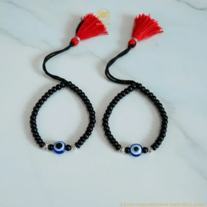 Divine Protection Wrapped in Artistic Elegance. Gift your little one the ultimate symbol of safety and style with the KJJ Signature Baby Nazariya. This premium set features a striking central blue evil eye charm, beautifully framed by 2.5mm chevron-textured imported silver balls. The intricate "V" patterns on the silver beads catch the light with every tiny movement, providing a sophisticated and luxurious sparkle that honors the sacred "Kala Dhaga" tradition. Handcrafted with certified 92.5 Sterling Silver, these bracelets are meticulously hand-strung with high-quality black beads on a soft, durable cord. Designed to be as gentle as a mother’s touch, this set ensures your baby remains shielded from negative energy while looking their absolute best. Key Highlights: Exclusive Chevron Texture: Features precision-crafted 2.5mm silver balls with an imported chevron-engraved finish for a high-end, multi-faceted shine. Certified 92.5 Purity: Every silver bead and accent is made from guaranteed high-purity sterling silver, making it hypoallergenic and safe for sensitive infant skin. Central Evil Eye Protection: Adorned with a vibrant blue evil eye charm at the center, traditionally used to ward off negativity and bring good fortune. Adjustable & Comfortable: Fitted on a soft black thread with a signature red and black tassel, allowing for a perfect, adjustable fit as your baby grows. A Meaningful Heirloom Gift: The perfect choice for Baby Showers, Naming Ceremonies, or as a protective first accessory for newborns. Product Specifications: Brand: Kanhaji Jewels (KJJ) Material: 92.5 Sterling Silver & High-Grade Black Protective Beads Silver Ball Size: 2.5mm (Imported Chevron-Cut) Category: Infant Protection Jewelry / Nazariya Quantity: 1 Pair (Set of 2)