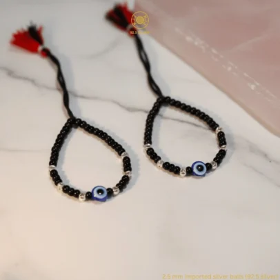 Artisan Sparkle for Sacred Security. Give your little one the ultimate symbol of safety and style with the KJJ Elite Baby Nazariya. This premium protective set features a vibrant central blue evil eye charm, beautifully accented by 2.5mm chevron-textured imported silver balls. The delicate "V" patterns engraved on the silver beads catch the light brilliantly, offering a sophisticated and modern twist on the traditional "Kala Dhaga". Handcrafted with certified 92.5 Sterling Silver, these bracelets are meticulously hand-strung on a soft, durable black cord with polished black beads to ward off negative energy. Designed specifically for sensitive infant skin, this set ensures your baby remains protected with high-quality materials and a touch of luxury. Key Highlights: 92.5 Sterling Silver Purity: Every silver bead and accent is made from certified high-purity silver, making it safe, hypoallergenic, and long-lasting for newborns. 2.5mm Chevron-Textured Beads: Features premium imported silver balls with an artistic faceted texture that provides more brilliance than standard smooth beads. Sacred Evil Eye Protection: Adorned with a striking blue evil eye charm at the center, traditionally used to shield children from negativity and bring peace. Adjustable Artisan Cord: Fitted on a soft black thread with a signature red and black tassel, providing a traditional look and a secure, adjustable fit as your baby grows. A Meaningful Blessing: An ideal choice for "Namkaran" (naming ceremonies), first birthdays, or a protective first accessory for a new arrival. Product Specifications: Brand: Kanhaji Jewels (KJJ) Material: 92.5 Sterling Silver & High-Grade Protective Beads Silver Ball Texture: 2.5mm Chevron-Cut (Imported) Category: Infant Protection Jewelry / Nazariya Quantity: 1 Pair (Set of 2)